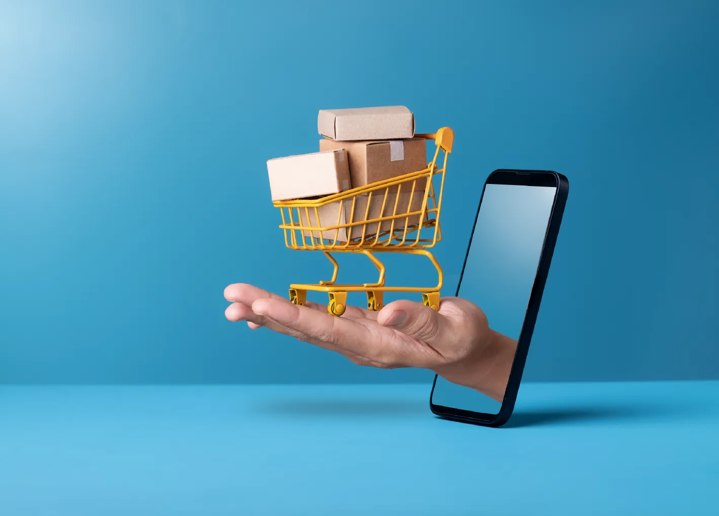 Mini shopping cart with cardboard boxes on hand through smartphone on blue background
