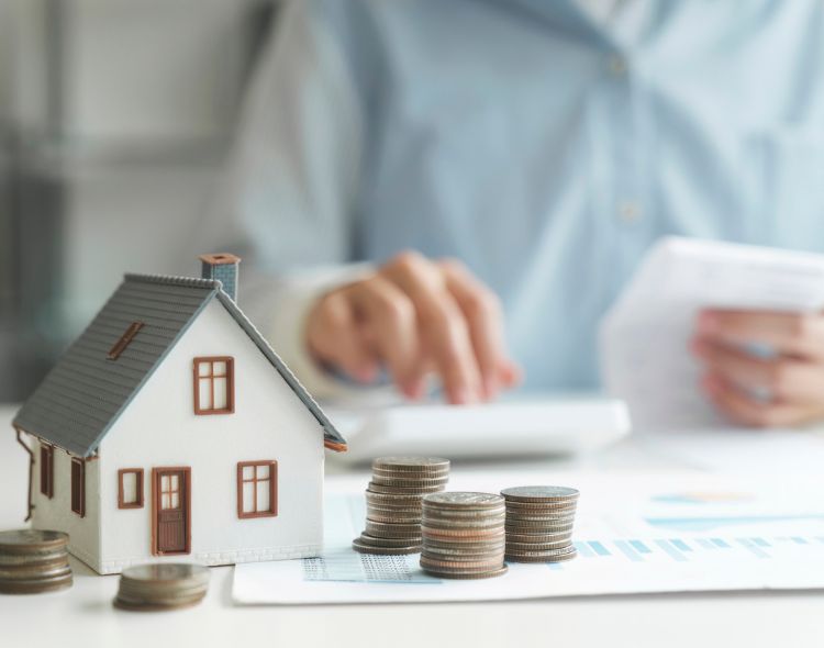 Small model house on desk with person in background looking at papers and coins in the foreground